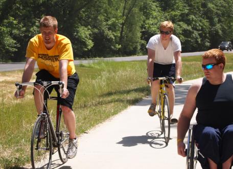 Example of inclusive recreation: Three young men, two on bikes and one wheelchair user, on a paved trail together.
